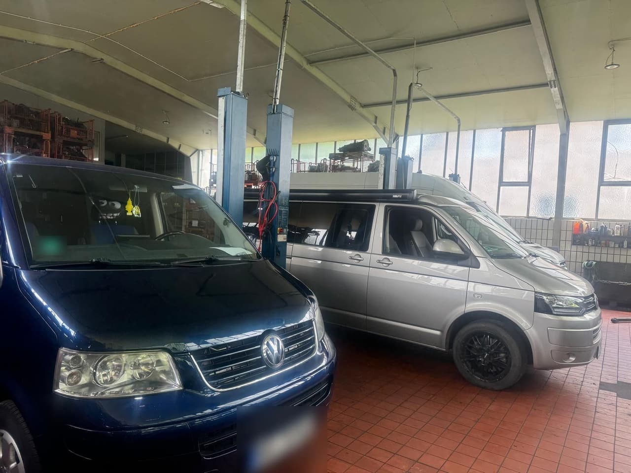 Zwei VW T5 in der Werkstatthalle, vorne ein blauer T5 vor dem Facelift, dahinter ein silberner T5 mit Markise