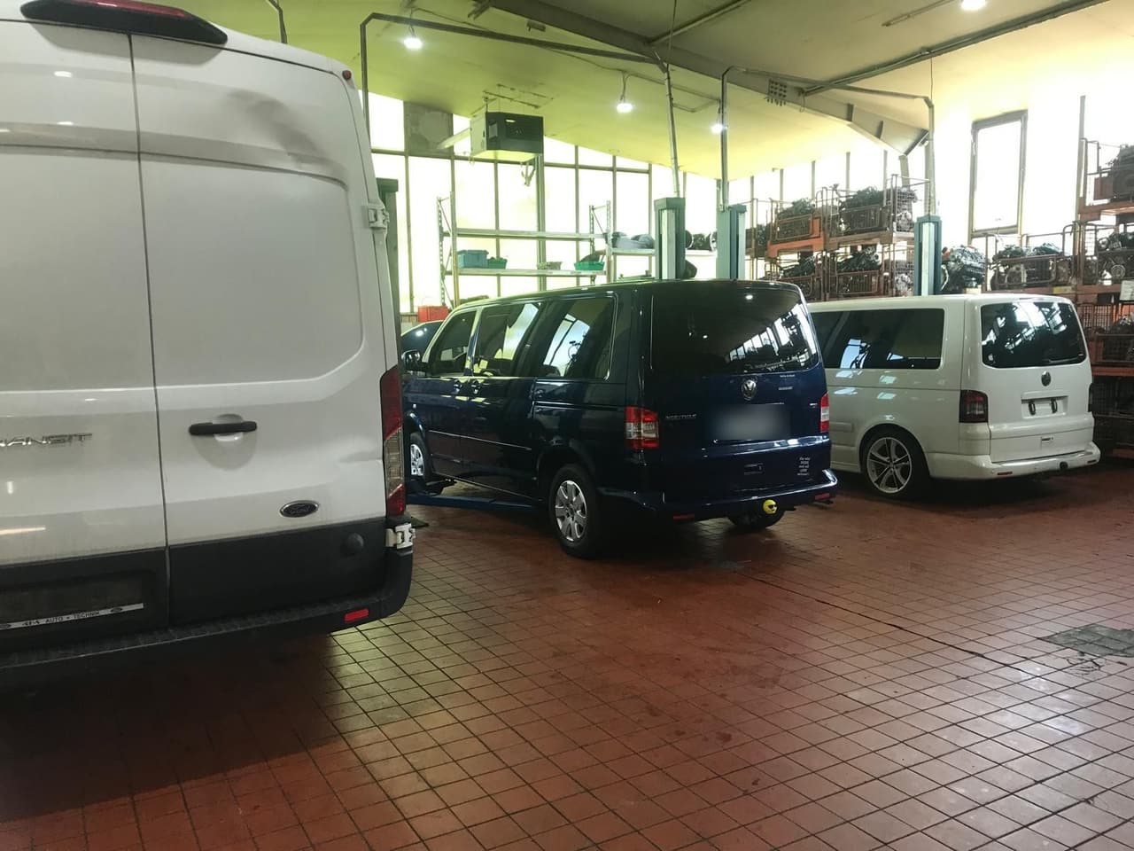Werkstatthalle mit Ford Transit Kastenwagen von hinten und zwei VW T5, im Hintergrund Regale mit Motoren
